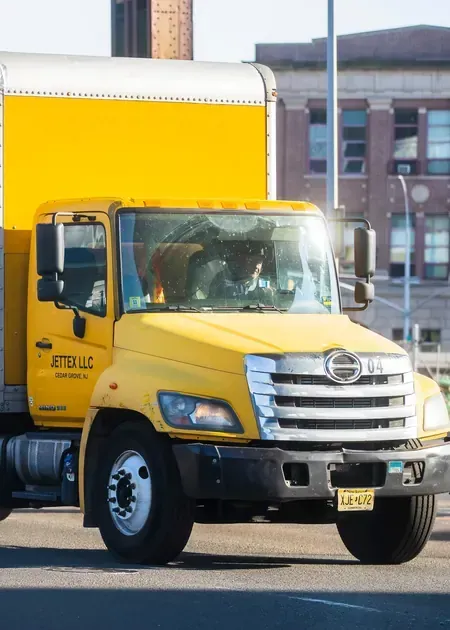 Yellow box truck driving on a city street in front of brick buildings.