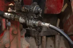 Close-up of a dirty metal hydraulic hose fitting leaking black fluid on red machinery.