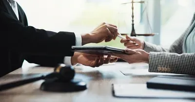 Person signs document during business handshake across a desk with a gavel and scales in the background