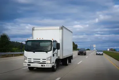 White box truck driving on a highway under a cloudy sky, with cars ahead.