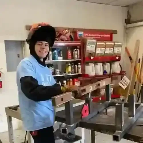 Person in a welding hood and blue apron smiles beside metal workpieces in a workshop.