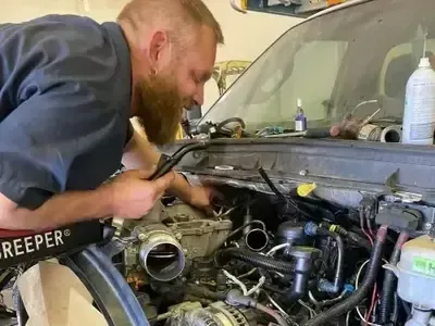 Mechanic leaning into a car engine bay, inspecting wiring under the hood.