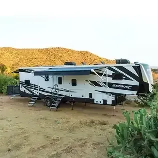 White RV camper parked on dirt near cacti, with desert hills in the background.