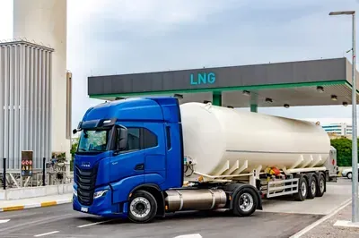 Blue LNG tanker truck at a fuel station under a canopy, with industrial tanks in the background
