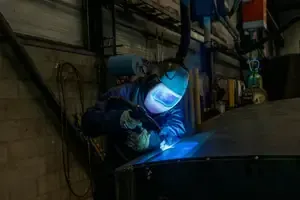 Welder wearing a face shield working on a glowing metal surface in a dark workshop