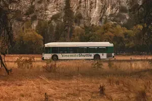 Yosemite shuttle bus in a grassy field with rocky cliffs and autumn trees in the background