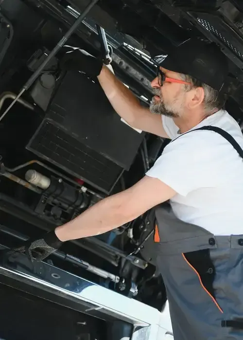 Mechanic working inside a car engine compartment, reaching upward with tools.