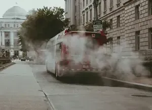 Red bus releasing steam on a city street beside stone buildings and a domed landmark.