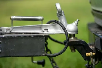 Close-up of a metal trailer hitch with wiring and a black box attached, against a blurred green background