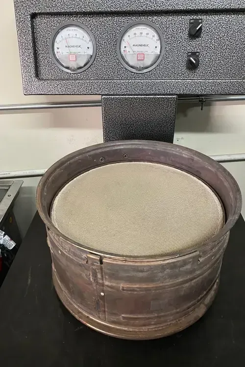 Round metal sieve filled with beige material on a black countertop beneath two gauges.