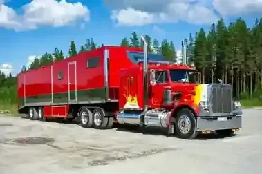 Red semi-truck with a bright red trailer parked on a gravel lot beside a pine forest under a blue sky.