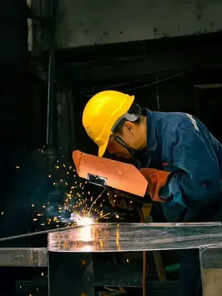 Worker in yellow hard hat and blue coveralls welding metal, sparks flying in a dark workshop