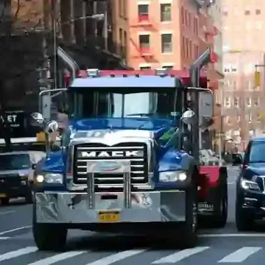 Blue Mack tow truck driving on a city street between buildings and cars