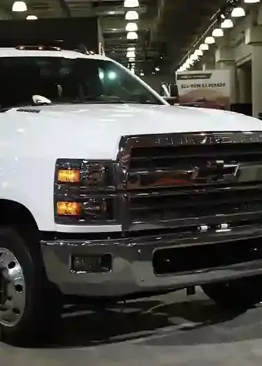 White pickup truck parked in an indoor garage, front view with chrome grille and headlights on.