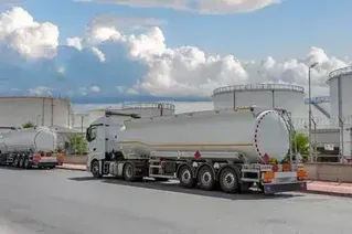 White fuel tanker truck parked on a road near industrial storage tanks under a cloudy sky