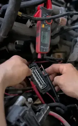 Hands using a red multimeter probe on a black electrical connector in an engine bay.