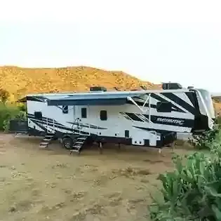Large white and black fifth-wheel RV parked on dirt in a desert campsite with hills behind