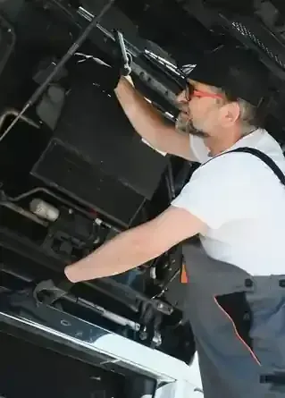 Person in a dark workspace reaching up to adjust equipment, wearing a white shirt and dark cap.