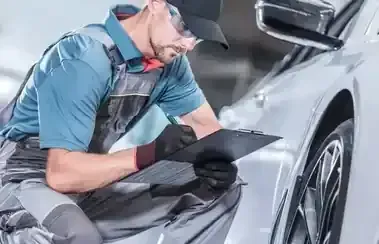 Mechanic crouching beside a silver car, inspecting a wheel with a tablet and gloves