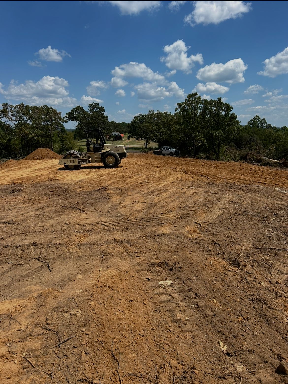 Construction site with dirt road grading, a bulldozer in the distance, and trees under a blue sky.