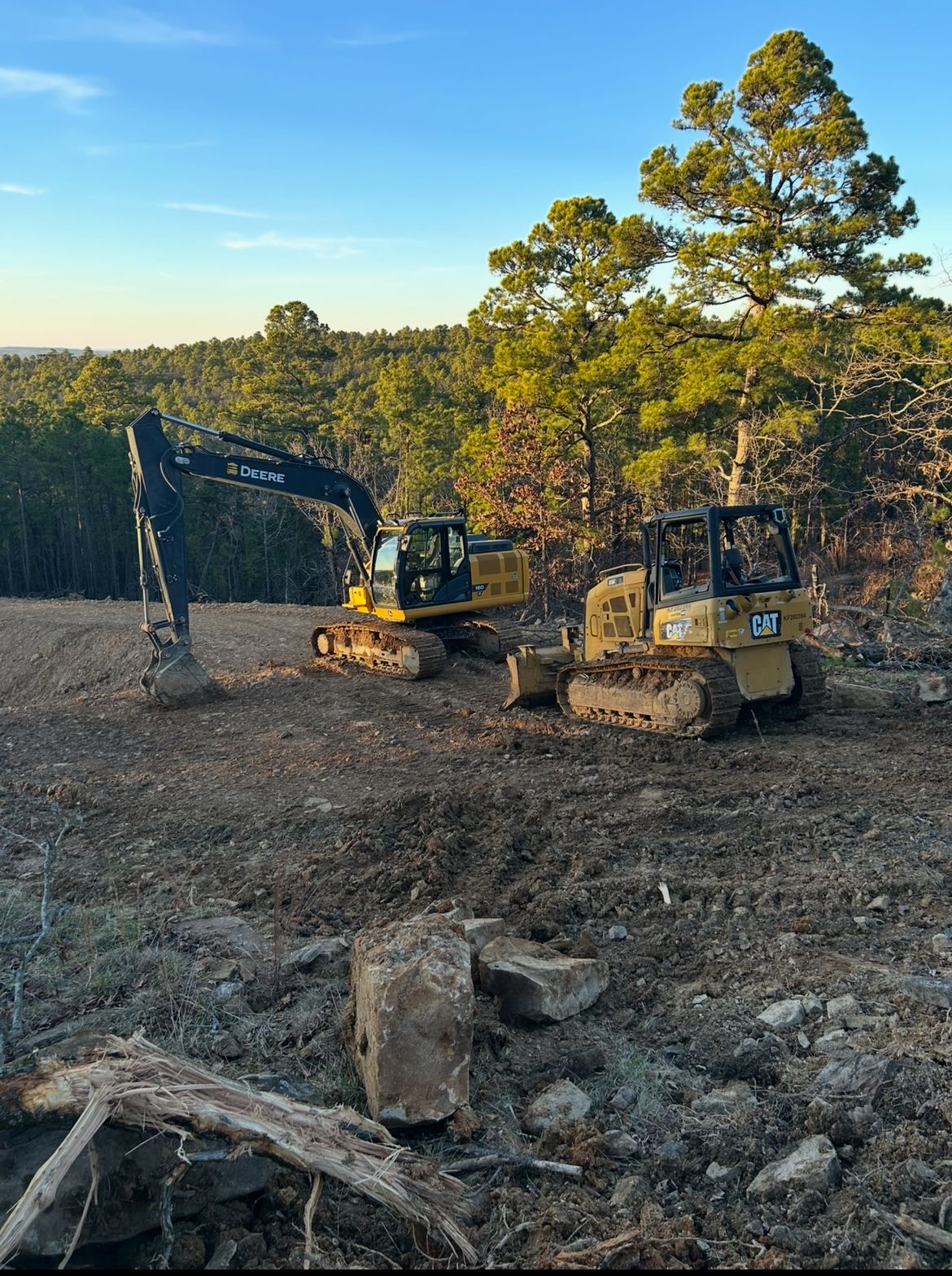 Two excavators clearing a dirt lot near tall trees at sunset
