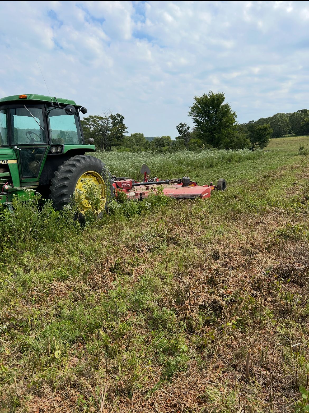 Green tractor mowing a grassy field with a red implement under a cloudy sky