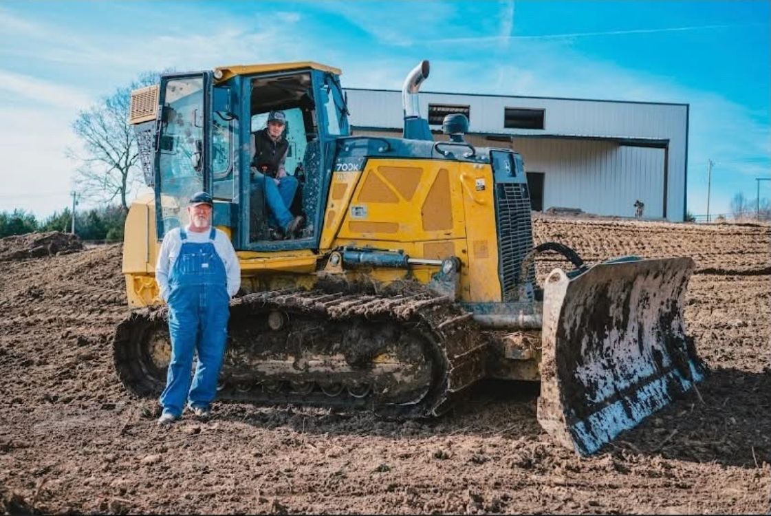 Yellow bulldozer with muddy blade beside a person in blue coveralls on a dirt site
