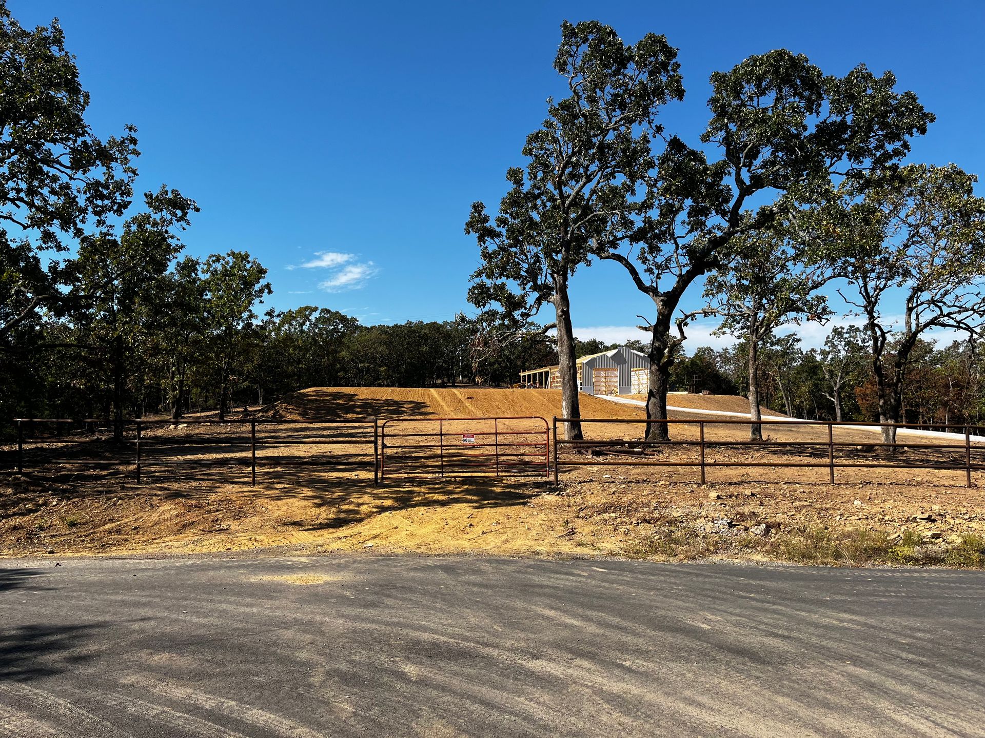 Dry rural roadside with a fenced field, trees, and clear blue sky