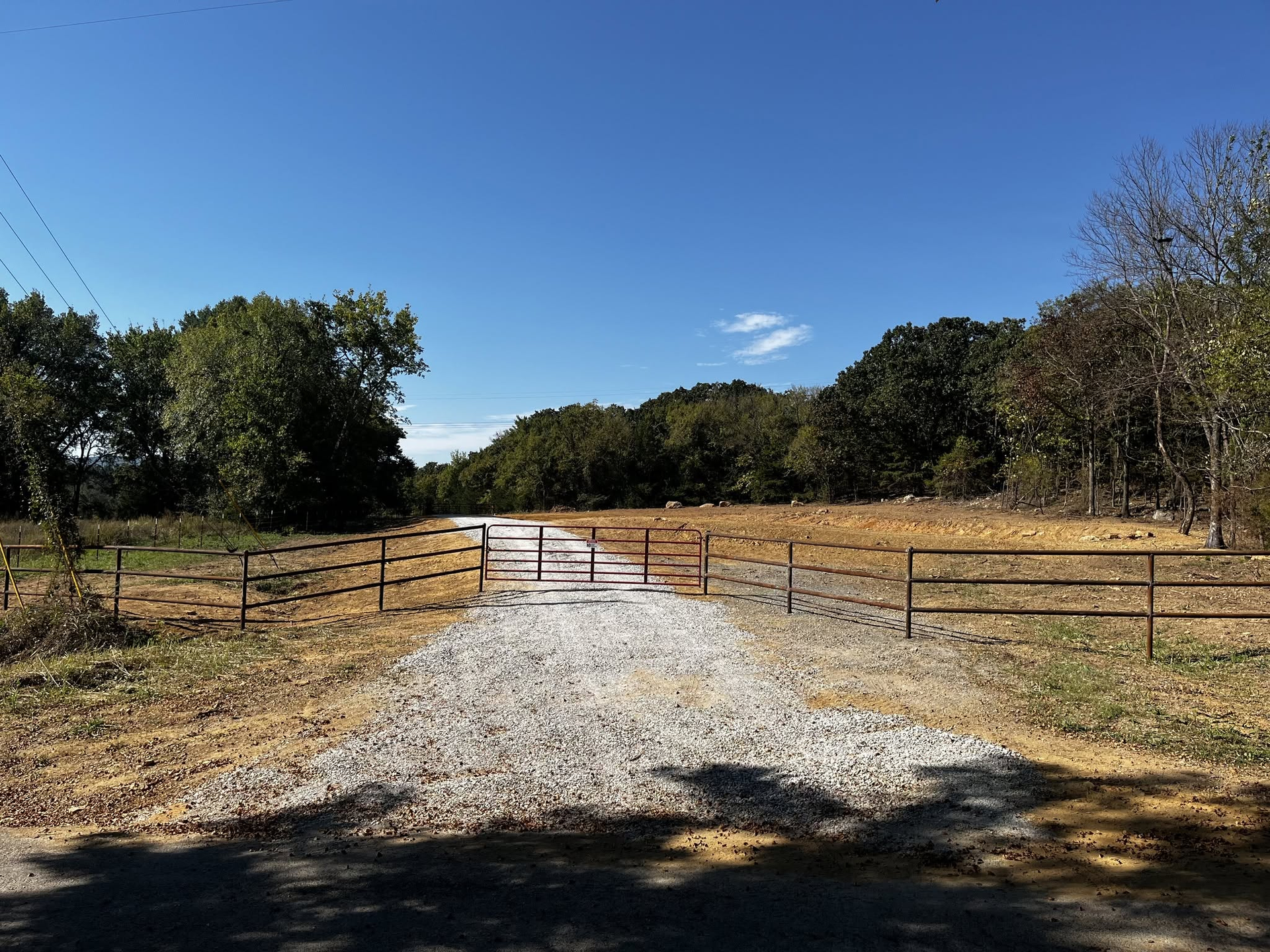 Gravel farm road leading to a metal gate, with dry grass, trees, and a clear blue sky.