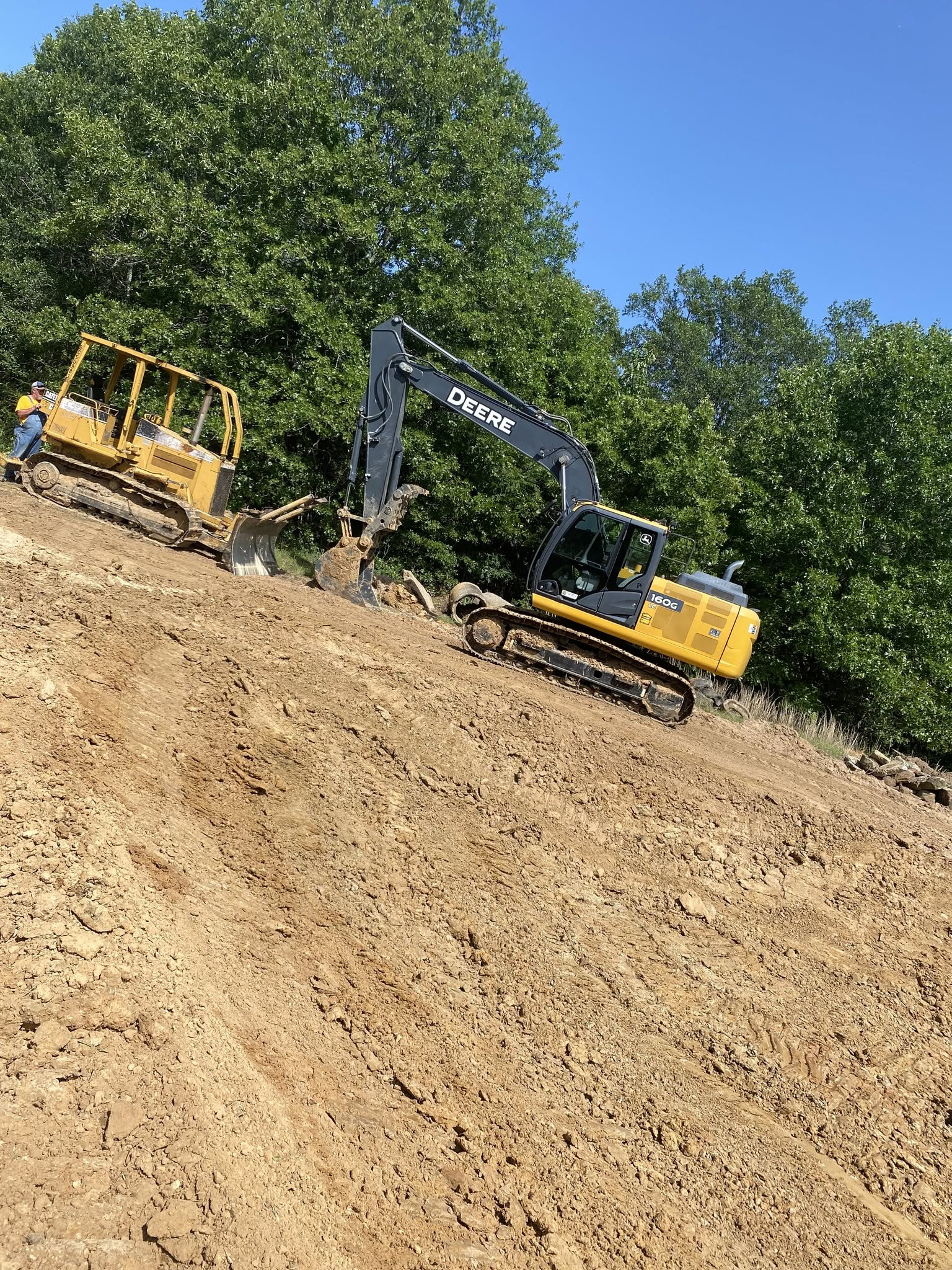 Yellow excavator and bulldozer working on a steep dirt slope beside trees under a blue sky