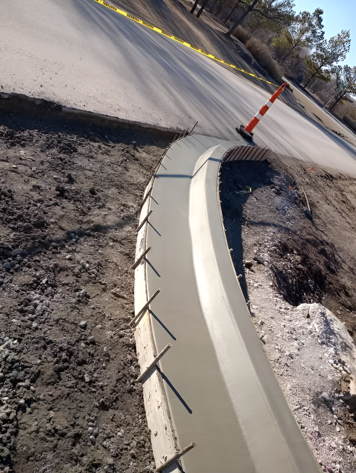 Road curb under construction beside a paved street with orange traffic cone and exposed dirt.