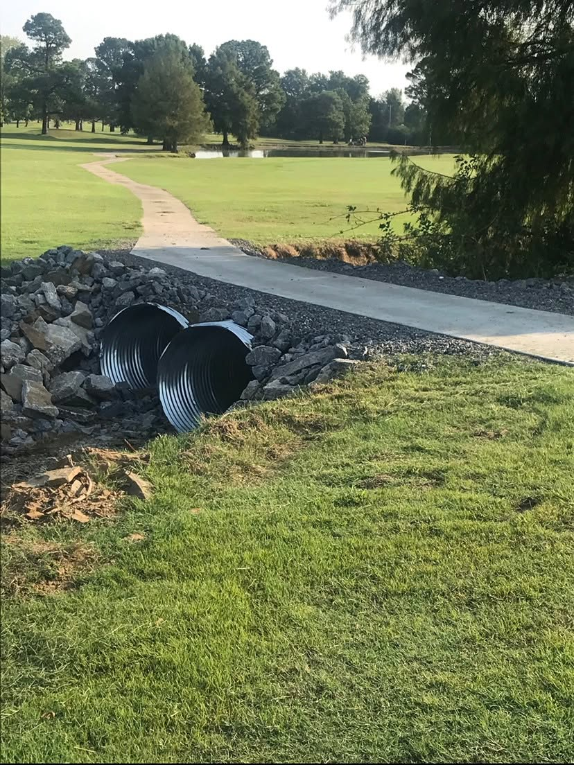 Storm drain culvert under a park path beside a grassy field and trees