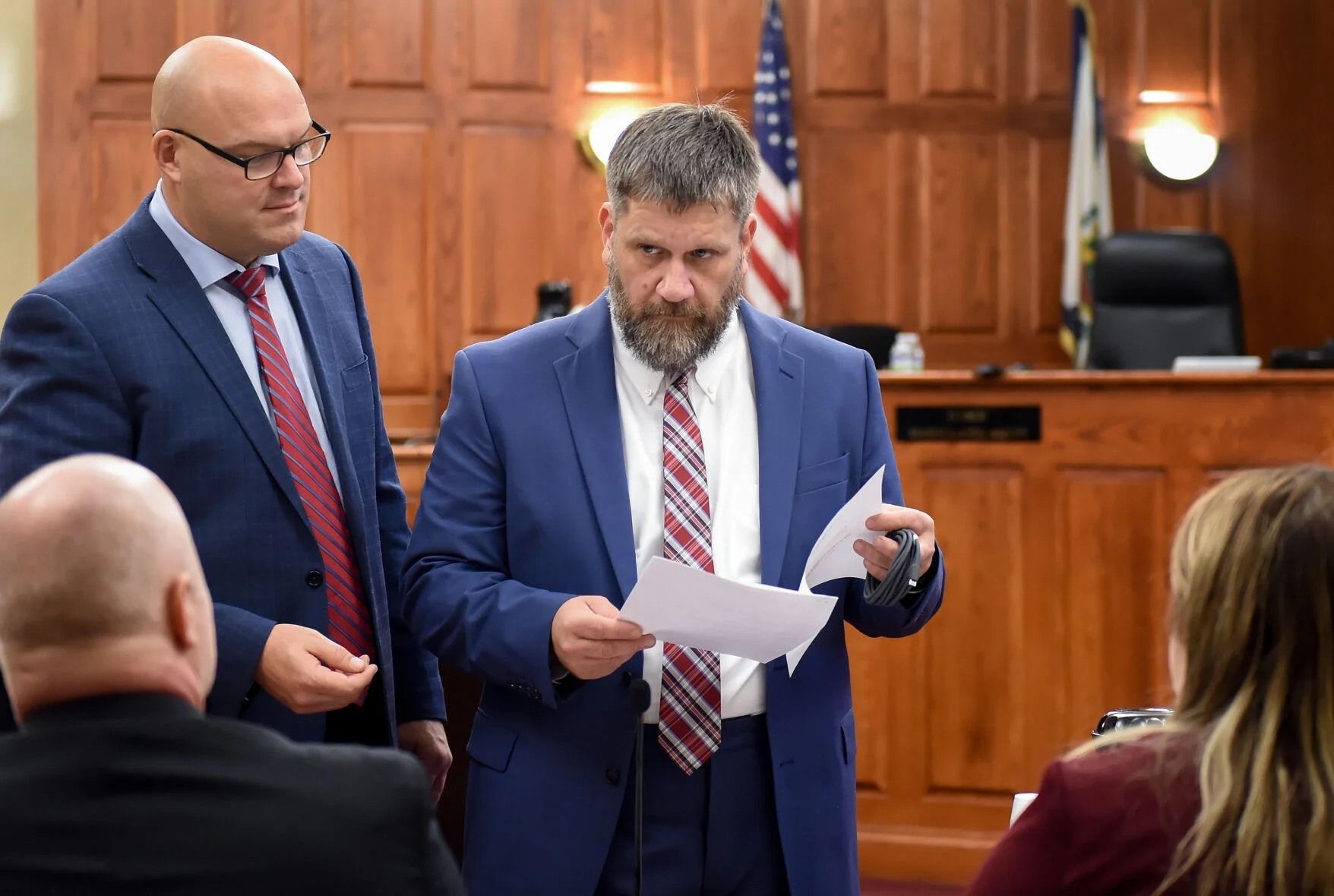 Two men in suits review a document in a wood-paneled courtroom.