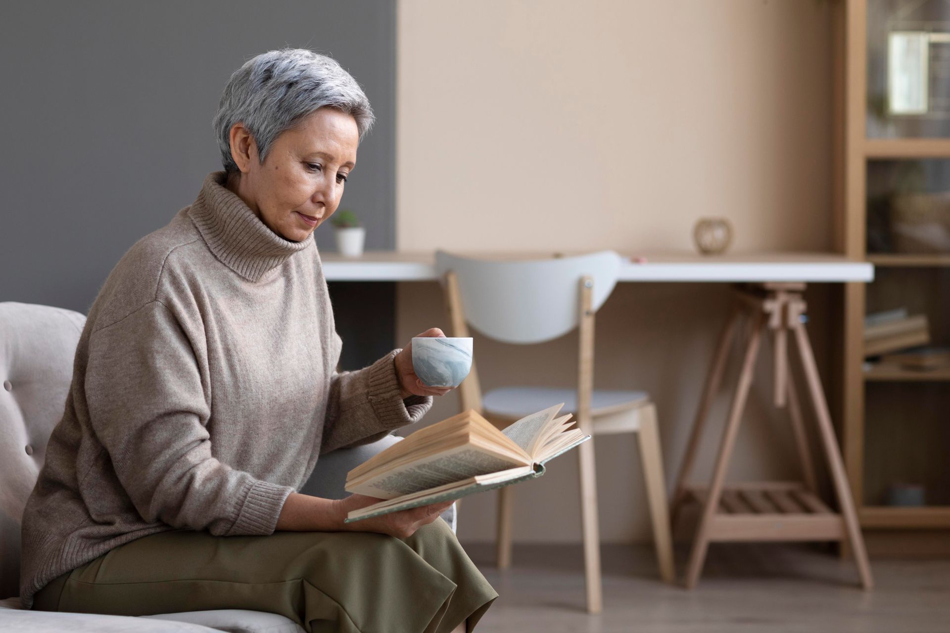 An elderly woman is sitting on a couch reading a book and drinking coffee.