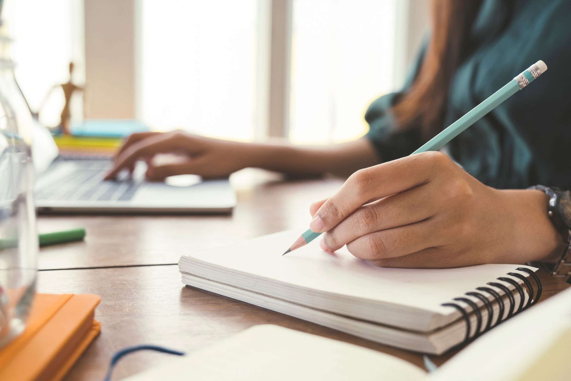 Person writing in a notebook with a pencil, laptop in the background. Wooden desk, bright daylight.
