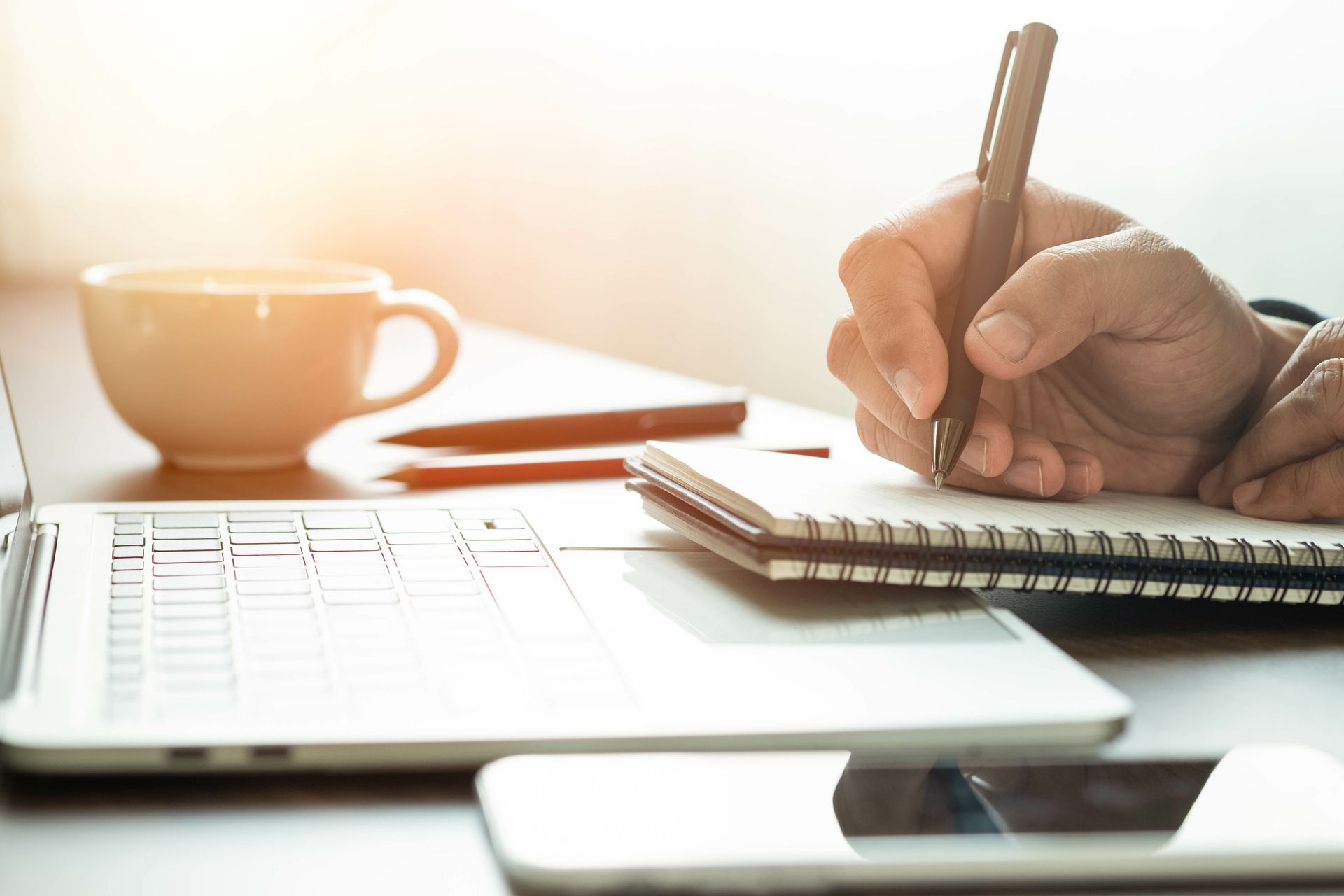 Hand writing in a notebook next to a laptop, phone, and mug on a desk, bathed in sunlight.