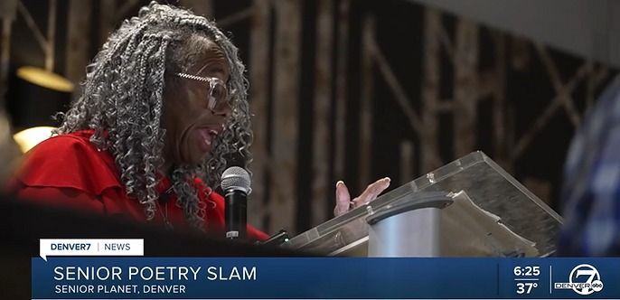 A woman with gray dreadlocks reads at a podium. The screen says