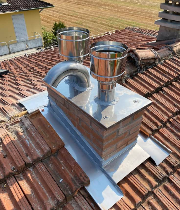 Camino sul tetto con due comignoli metallici e una lamiera argentata su tegole di terracotta rossa.