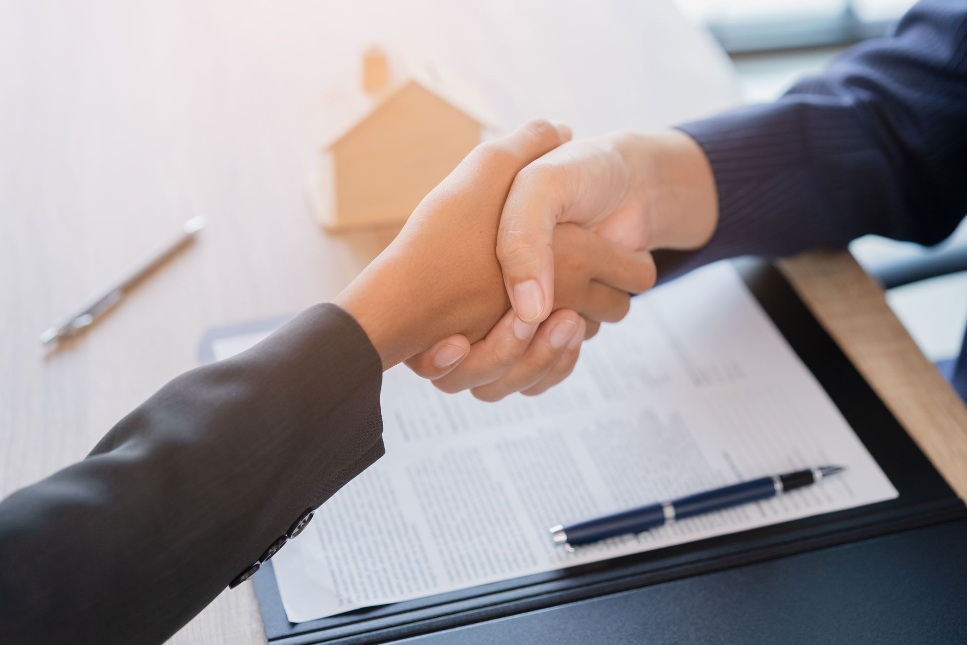 Two people shaking hands over a contract, miniature house and pen on a desk.