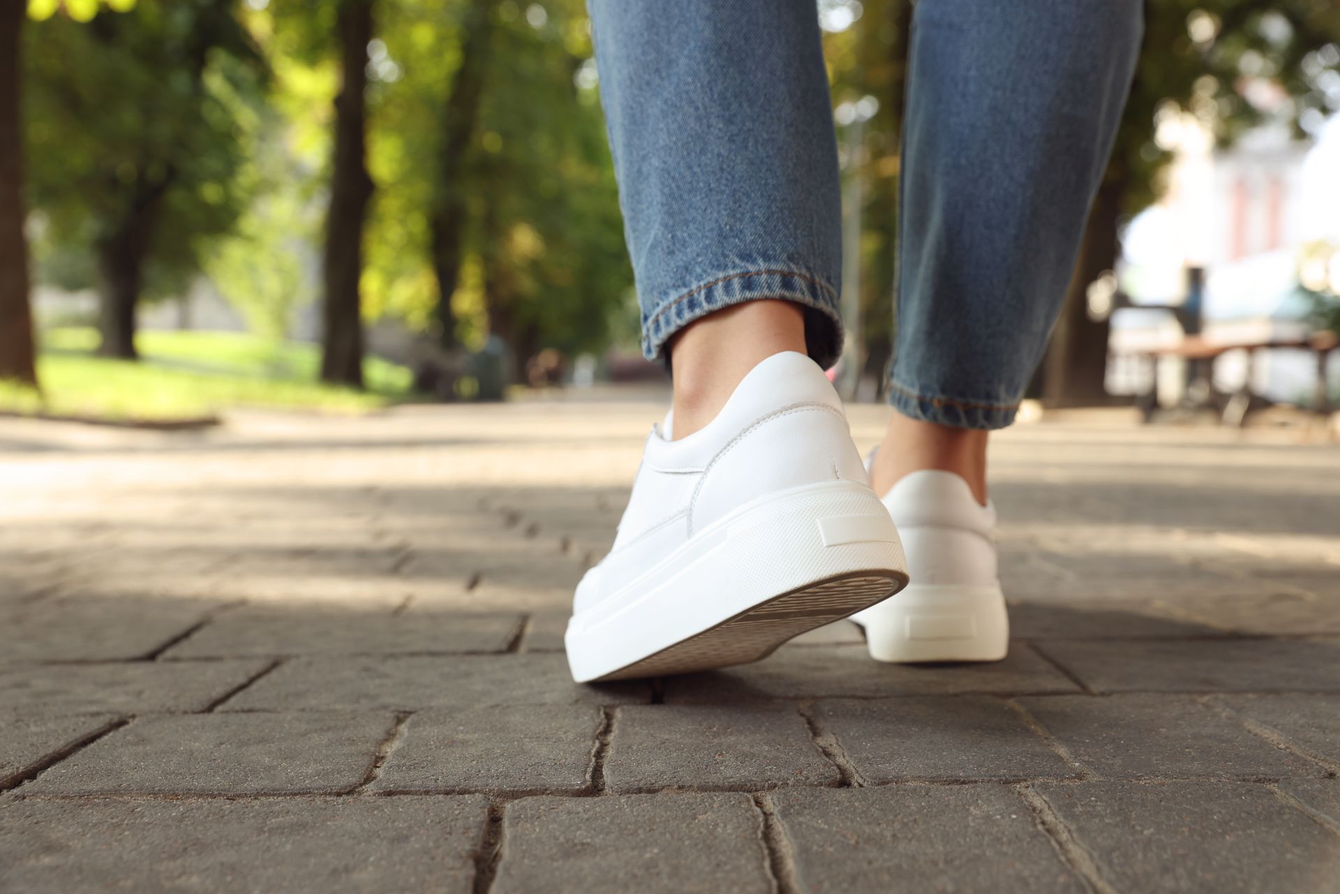 Person in blue jeans and white sneakers walking on a brick path outdoors.