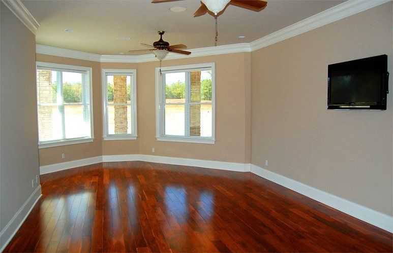 An empty room with polished hardwood floors, beige walls, white trim, a bay window with a view, and a wall-mounted TV.