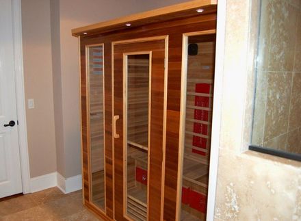 A wooden indoor sauna with a glass door, built-in lights, and red heater panels, situated in a tiled room.