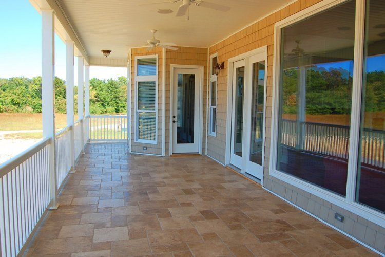 A covered outdoor patio featuring tan tile flooring, white railings, and a view of a grassy landscape.