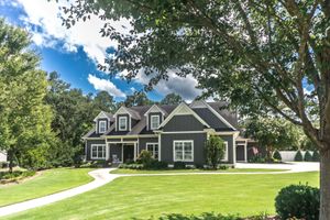 A dark gray, multi-story house with white trim and three dormer windows, surrounded by a green lawn and large trees.
