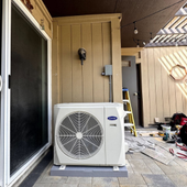 Outdoor HVAC unit beside a house wall, with a ladder and tools on a covered patio during installation