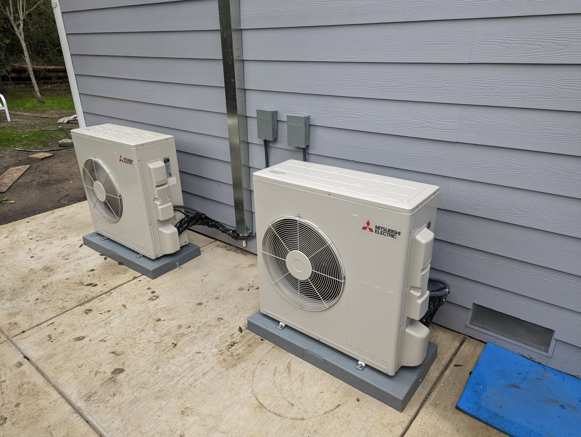 Two outdoor air-conditioning units beside a gray house on a concrete slab.