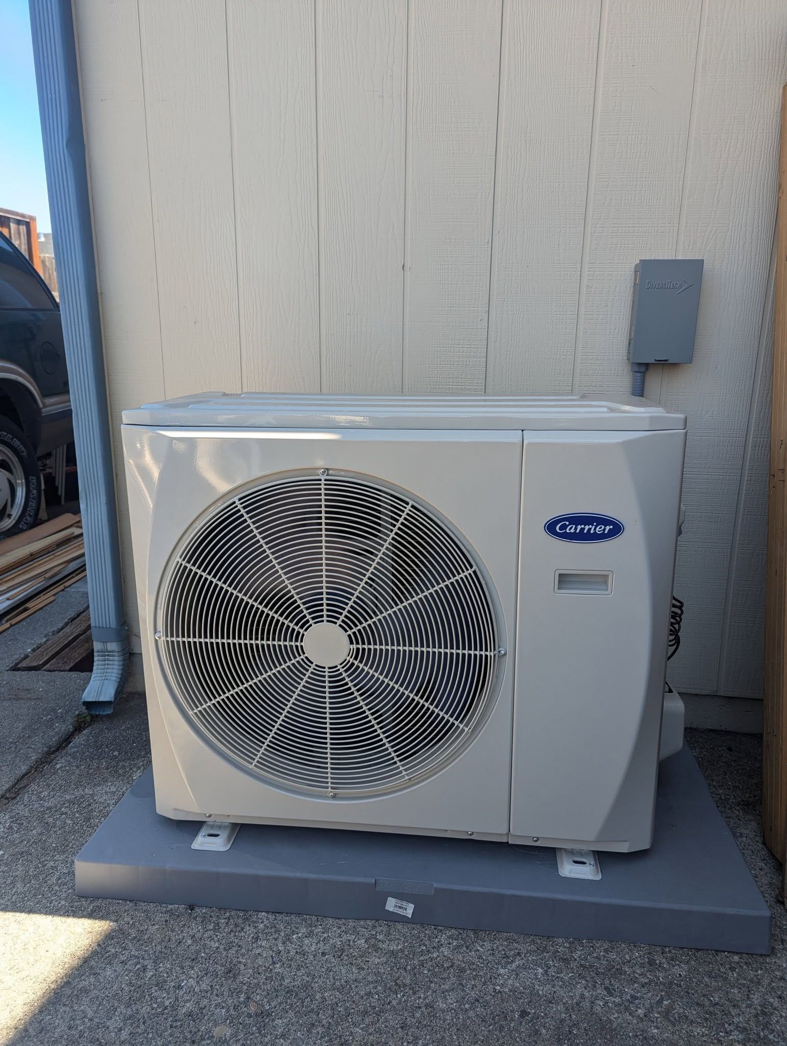 Outdoor HVAC condenser unit beside a building wall on a gravel pad, with a protective fan grille and electrical box.