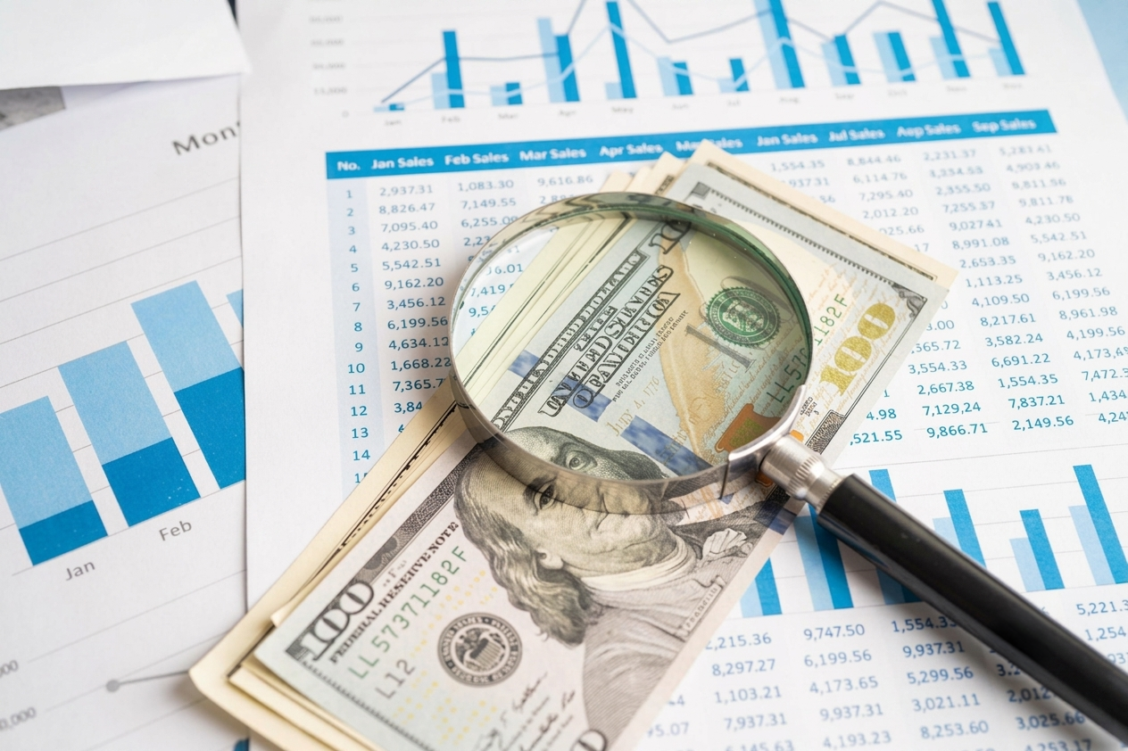 Magnifying glass over dollar bills and financial charts on a desk