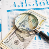 Magnifying glass over U.S. hundred-dollar bills and financial charts