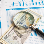 Magnifying glass over U.S. hundred-dollar bills and financial charts